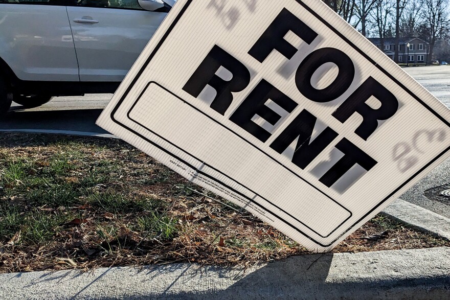 A white "For Rent" sign with black letters stands propped up sideways on the sidewalk 