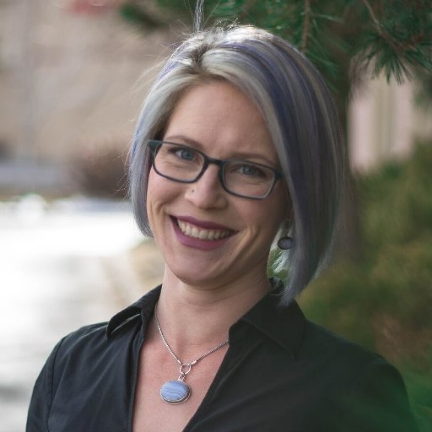 Sarah Peters is looking at the camera and smiling. She is wearing a black shit and has purple streaks in her hair. 