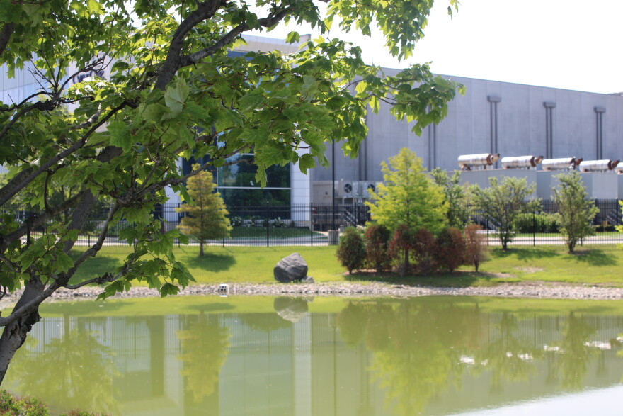 A photo of a pond in front of a data center fenced off behind it, a tree is close to the screen. 