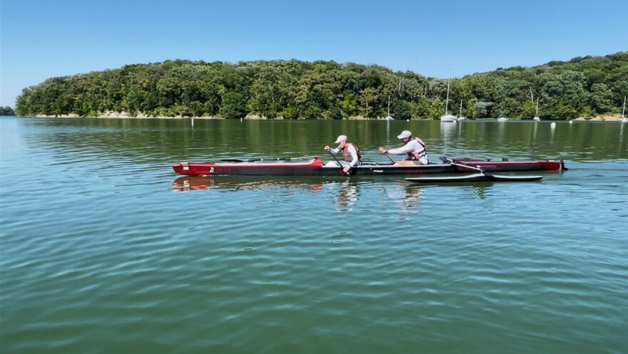 Side view of two people in a long, narrow boat paddle on a body of water.