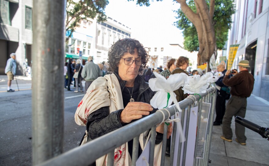 Rabbi Cat Zavis ties a white paper flower to a metal fence outside ICE in San Francisco. Zavis is a woman with curly chin-length gray hair wearing a yarmulke, square glasses, a silver necklace and a hand-painted robe over a black jacket. 