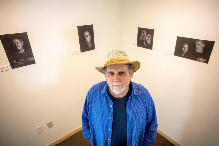 A man in a blue long-sleeved shirt over a dark blue t-shirt wearing a straw hat looks up toward the camera. The man has a short, white beard and stands in front of two walls painted off-white displaying black and white photographs.