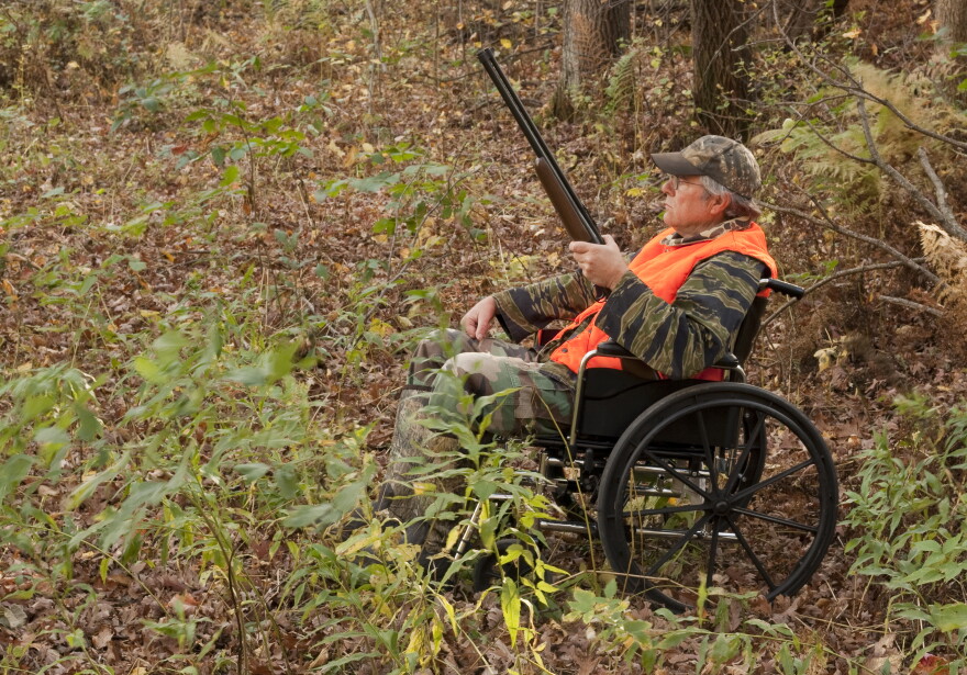 A hunter using a folding-frame wheelchair sits in a wooded area during fall. He is holding a shotgun and wearing camouflage clothing with a bright orange safety vest and camouflage cap.