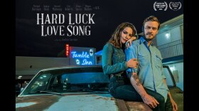 The movie poster for “Hard Luck Love Song.” A man is leaning against a car. A woman is sitting on the hood of the car while wrapping her legs around the man. They are looking toward the camera with content expressions.