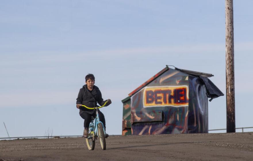 A boy bicycles through town on Wednesday, April 2, in Bethel, Alaska. The Lower Kuskokwim School District, headquartered in Bethel, has shipping containers full of contaminated materials that have been there for years, waiting to be barged out. (Photo by Rashah McChesney/Alaska’s Energy Desk)