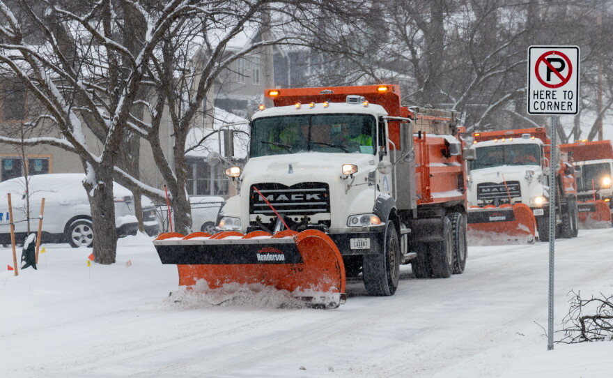 A line of three snow plows on a snow-covered city street.