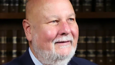 A round-faced, bald caucasian man with a grey mustache and trimmed beard, in front of a wall of books, looking off-camera and smiling slightly.