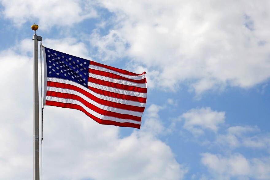 A red, white and blue American Flag is blowing in the wind in front of a cloudy blue sky.