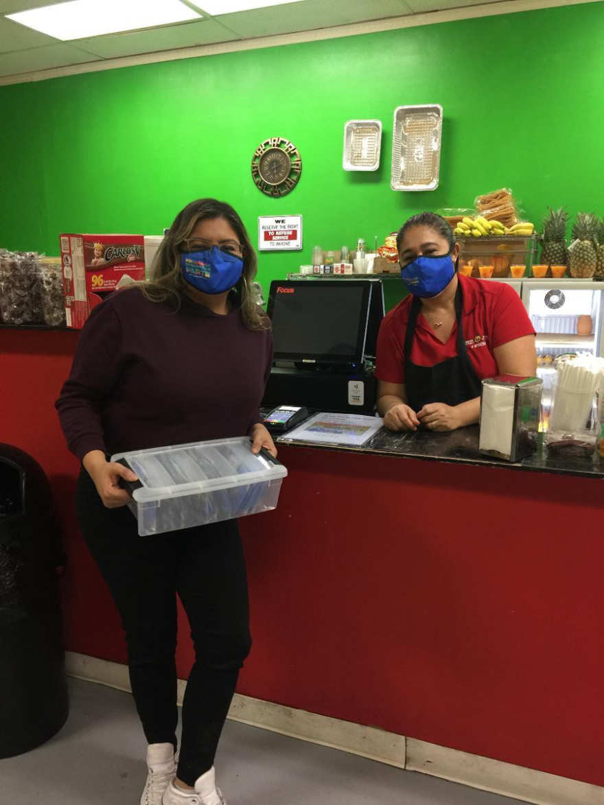 Ivet is standing near an employee at a local business. Both are women and are wearing facial masks. Ivet is also holding a box of facial masks.