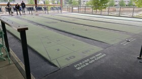 Five green and blue shuffleboard courts with some people playing the game beneath a gazebo. 