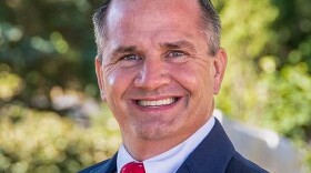 A man wearing a dark suit, white shirt, and red tie smiles at the camera. He has short, neatly styled hair and is standing outdoors with soft, green foliage blurred in the background.