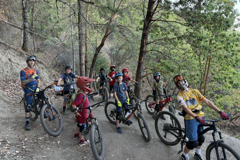A group of children wearing helmets stand with mountain bikes on a wooded dirt trail during an Ashland Devo youth ride.