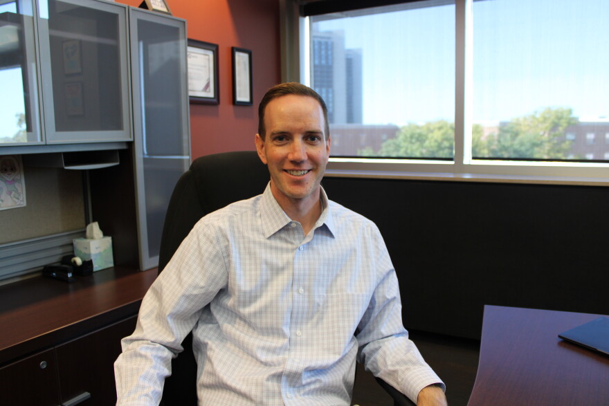 A man in a button down shirt sits at an executive desk in an office and smiles.