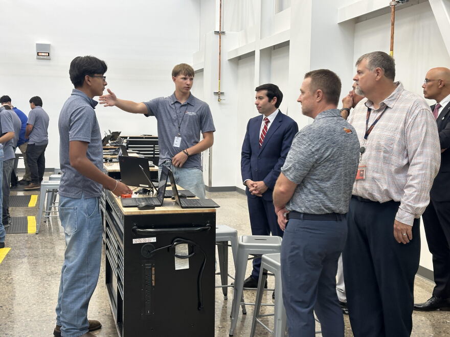 A group of professionally dressed, middle-aged men listen to two high-school-aged boys talk about what they're learning in a warehouse-style classroom.