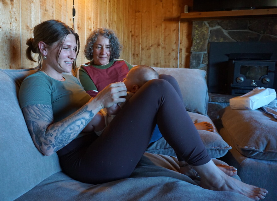 A woman with tattoos on her right arm holds a small baby in her lap on a grey couch. Another woman sits next to her, gazing at the baby.