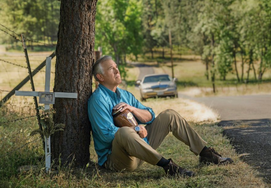 Bruce Campbell in a still from "Ernie & Emma." He's leaning against a tree, sitting down and holding an urn. To the left is a roadside cross.