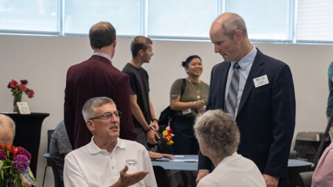 A man sitting at a banquet table gestures at a man standing near him. 
