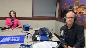 Two smiling people wearing headphones sit at a table in a radio talk studio: a woman back left has short light brown hair and is wearing a pink top; a man front right has very short dark hair and is wearing glasses, a black button-down shirt, and jeans