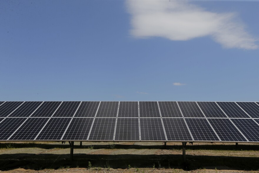 A wide shot of a ground mounted solar array. The panels run across the bottom half of the image. A bright blue sky and a white cloud are visible at the top. 