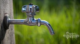 A close-up phot of a silver metal water faucet in focus in front of a blurred green grass background.