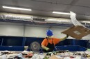 A recycling facility employee holds a large piece of cardboard in their hand while looking down at the streamline of recyclable materials.