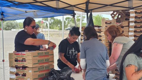People crowd under a canopy with stacks of boxes of produce.