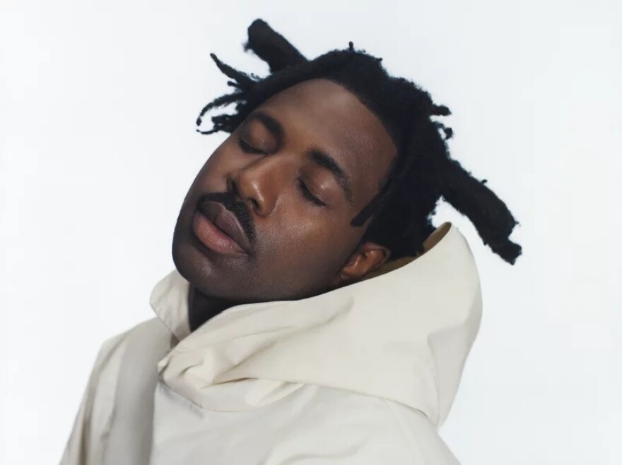 A man wearing a white hoodie tilts his head back with his eyes closed while standing in front of an all-white background.