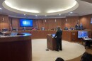 A man in all green stands in front of a dais of county commissioners