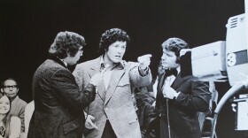  man in 1970s stands flanked by two other men and speaks into a television camera