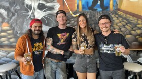 Three men and a woman stand in a group in front of a large mural holding pints of beer while smiling at the camera.