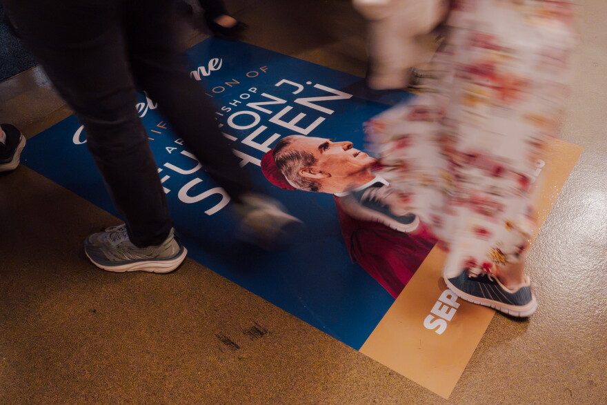 People walk over a floor graphic depicting the late Archbishop Fulton J. Sheen as they enter an expo hall ahead of his beatification Mass on Thursday.