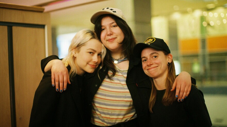 Three women smile at the camera, with one in the middle putting her arms around the two on either side of her.