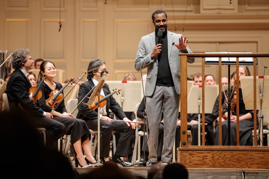 Carlos Simon, wearing a light grey suit, stands at the front of the stage at Symphony Hall. He holds a microphone and is addressing the audience, gesturing with his other hand.