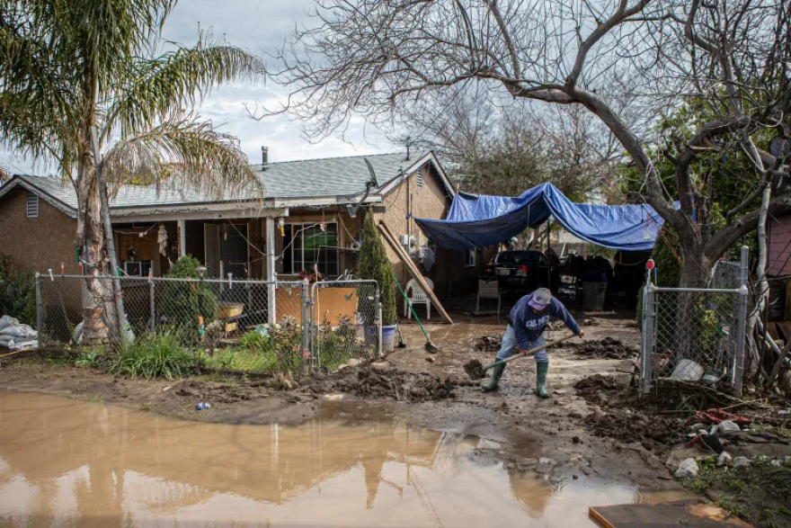 A flooded street leading to a driveway where a man shovels mud off his driveway.