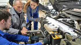 Two apprentices assist in working on a car engine.