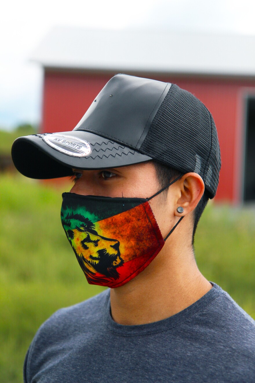 English: A photo of a person wearing a face mask, a baseball cap and a dark shirt, set against a blurred background of grass and a red building.
Español: La foto de una persona utilizando un cubreboca, una gorra de baseball y una camiseta oscura, sobre un fondo difuminado que revela césped y un edificio rojo.