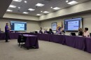 A man stands in front of a group assembled around a board room, presenting on a large screen. 