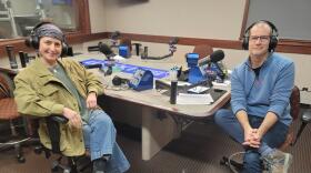 Two people wearing headphones sit at a table in a radio talk studio: a woman at left has a blue headscarf and is wearing a green jacket, green shirt, blue jeans and grey boots; a man at right has very short dark hair and is wearing glasses, a light blue quilted sweatshirt, blue jeans and brown boots.