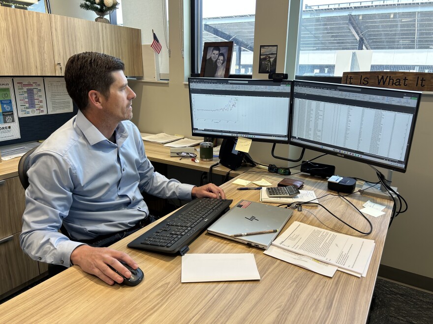 A man in a blue dress shirt sits at a desk in an office building and looks at two computer screens displaying spreadsheets of data about outdoor water usage. 
