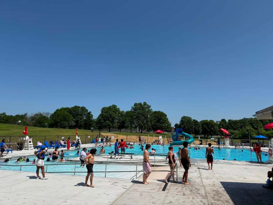 The picture shows dozens of people using the pool at the Knickerbacker Aquatic Center. A wide ramp descends into the pool. Lifeguard chairs and red umbrellas sit along all four sides of the pool. Trees and grass from the adjoining park are visible in the background. 