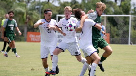 3 soccer players in white uniforms compete with a player in green for a loose ball