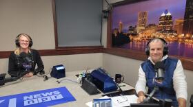 Two people wearing headphones sit at a table in a radio talk studio: a woman at left has long blonde hair and is wearing glasses and a long-sleeved black shirt; a man at right has short dark hair and is wearing a puffy navy vest over a white shirt.