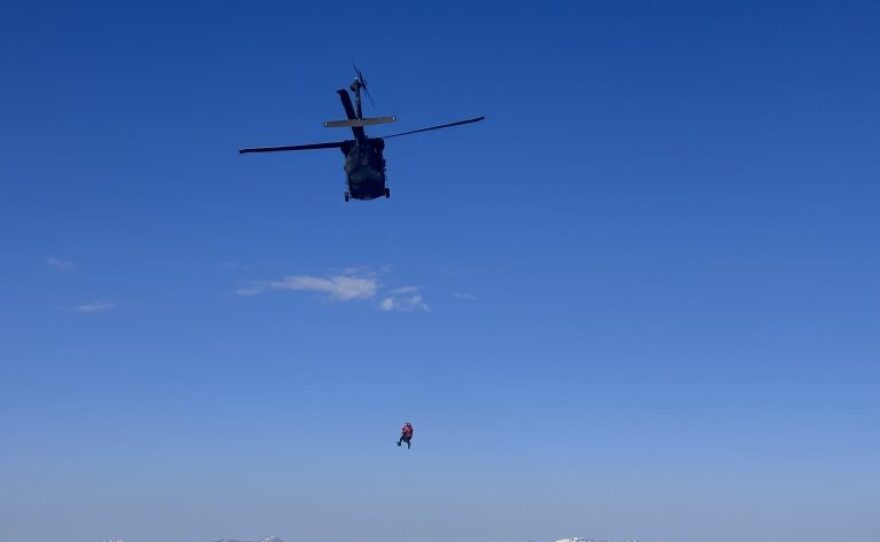 A helicopter flies above snowcapped peaks with a person attached to a line below.