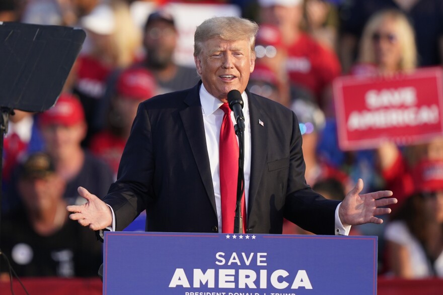 a man in a suit, white shirt and red tie speaks at a podium with a crowd behind him