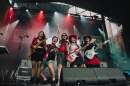 Fvie women in red and black outfits that look like a mashup between cowboy and goth pose on a stage for a photo.