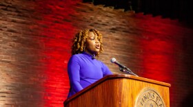 Karine Jean-Pierre stands at a wooden podium delivering a speech. She is wearing a blue dress, hoop earrings and curled hair. 