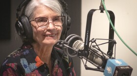 A woman sits inside a radio studio. She is smiling and talking at a microphone.