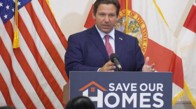Un hombre con traje azul habla desde un podio, frente a una bandera de Estados Unidos y una de Florida. El podio lleva un letrero que dice "SAVE OUR HOMES" (Salven nuestros hogares).