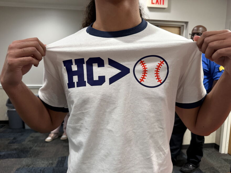 A man grabs the sides of his white shirt to showcase the text that reads "HC > Baseball," but the baseball is an icon, and not words.