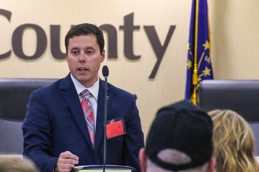 Alex Zimmerman stands in front of a lectern with a microphone on it, speaking to a crowd in front of him. Zimmerman is a White man with dark hair, wearing a suit.